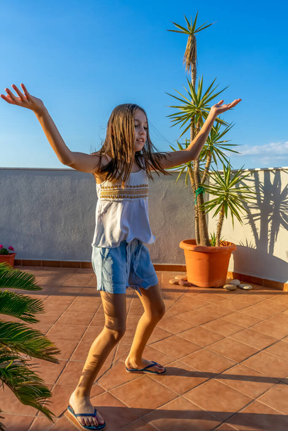 Linda niña vestida con una camiseta blanca bailando y saltando en la terraza de la casa al atardecer. Concepto divertido
 - Foto, imagen