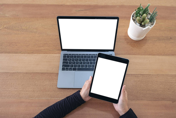Imagem de maquete de vista superior de mãos segurando um tablet preto e laptop com tela branca em branco na mesa de madeira no escritório
 - Foto, Imagem