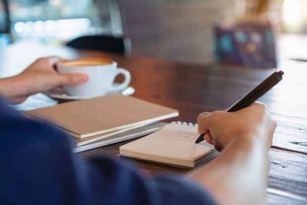 Closeup image of a woman holding and drinking hot coffee while writing on notebook on wooden table in cafe - 写真・画像