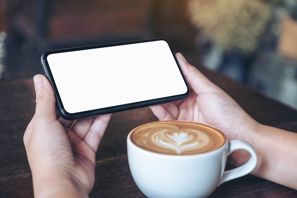 Образец изображения рук, держащих и использующих черный мобильный телефон с пустым экраном горизонтально для просмотра с чашкой кофе на деревянном столе
 - Фото, изображение