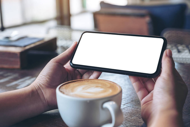 Wizerunek dłoni trzymających i korzystających z czarnego telefonu komórkowego z pustym ekranem poziomo do oglądania z filiżanką kawy na drewnianym stole - Zdjęcie, obraz