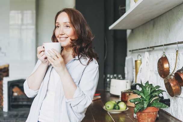 Ενήλικες χαμογελαστή μελαχρινή γυναίκα σε casual με φλιτζάνι τσάι στην κουζίνα στο σπίτι - Φωτογραφία, εικόνα
