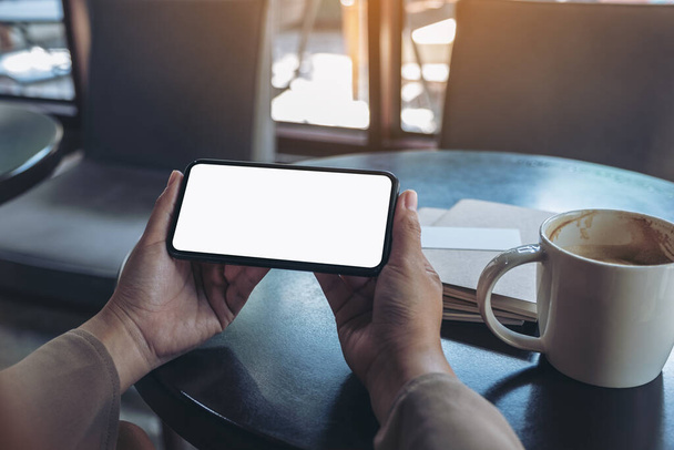 Mockup image of woman's hands holding and using a black mobile phone with blank screen horizontally for watching with notebook and coffee cup on wooden table - Photo, Image