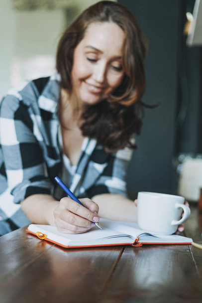 Adulto sorrindo mulher morena no casual fazendo notas no livro diário com xícara de chá na cozinha em casa
 - Foto, Imagem