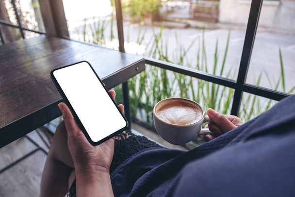 Imagen burlona de una mujer sosteniendo un teléfono móvil negro con pantalla en blanco mientras bebe café en la cafetería - Foto, imagen