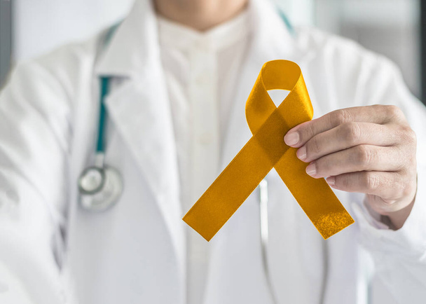 Conciencia del cáncer infantil con cinta de oro color de lazo simbólico en la mano del médico  - Foto, Imagen