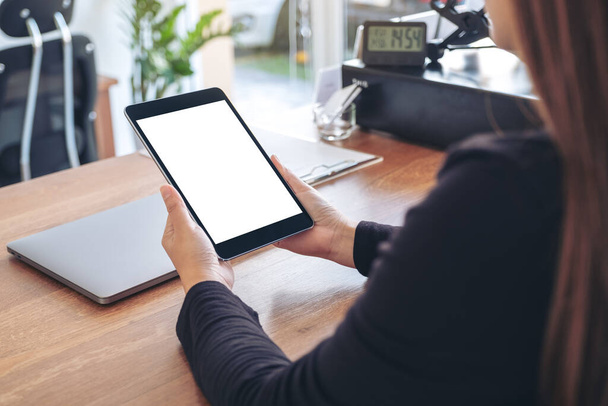Mockup image of a woman holding and using black tablet pc with blank white desktop screen with notebook on wooden table in office - Valokuva, kuva