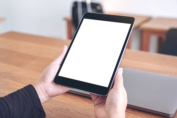 Image maquette des mains tenant et utilisant une tablette noire pc avec écran blanc vierge de bureau avec ordinateur portable sur une table en bois dans le bureau - Photo, image