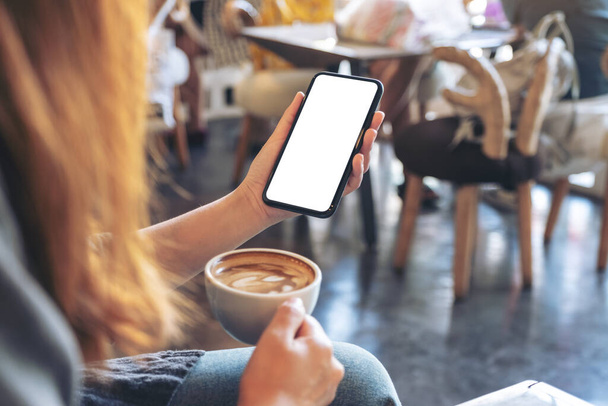Mockup obraz ženy držící bílý mobilní telefon s prázdnou obrazovkou při pití kávy v moderní kavárně - Fotografie, Obrázek