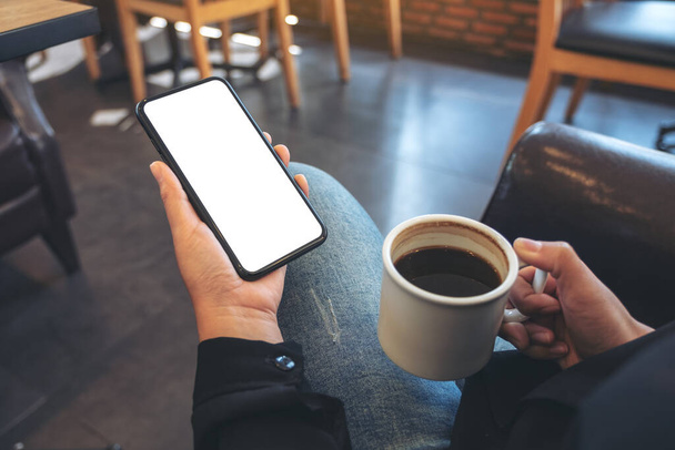 Mockup kuva kädet tilalla valkoinen matkapuhelin tyhjällä näytöllä juodessaan kahvia modernissa kahvilassa - Valokuva, kuva