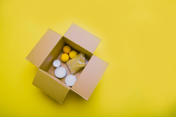 the main products for self-isolation in a box: cereals, buckwheat, fruit, canned food on a yellow background. home delivery. Assistance to population - Фото, изображение