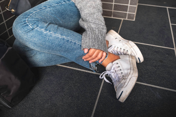 Detailní záběr ženy v džínových a bílých teniskách dotýkající se její nohy, když sedí na podlaze - Fotografie, Obrázek