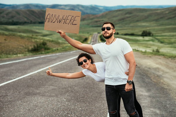 A couple hitchhiking. A man and a woman catch a car by the road. A young couple votes on the road. A man and a woman are hitchhiking. A couple hitch a ride on the road. Hitching a ride - Zdjęcie, obraz