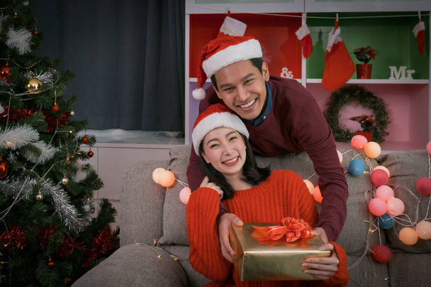 heureux couple charmant homme donner un cadeau ou des cadeaux à la femme, ils tenant cadeau et célébrer ensemble à la nuit de Noël dans la chambre qui décoré avec arbre de Noël pour le jour du festival de Noël asiatique - Photo, image