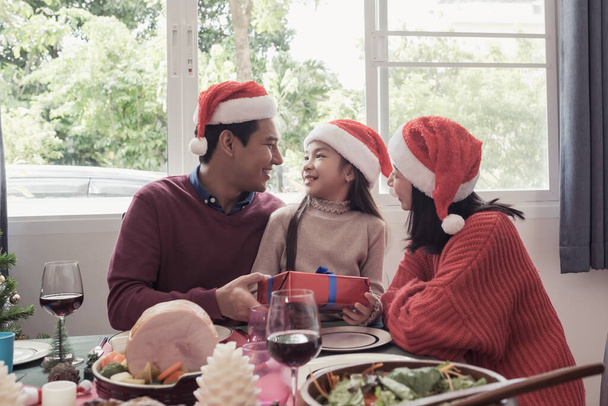 Aziatisch familie dochter houden geschenk met vader en moeder op kerstdag, ze zijn gelukkig en samen eten in de eetkamer die versierd met kerst thema - Foto, afbeelding