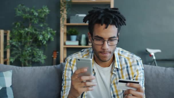Mixed race man shopping online making transaction with credit card and smartphone smiling at home - Imágenes, Vídeo