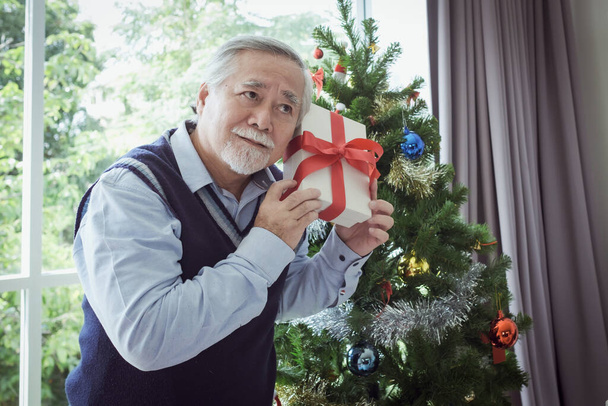 ギフトボックスを持った年配の男性の肖像画と笑顔でクリスマスツリーの前に立つ - 写真・画像