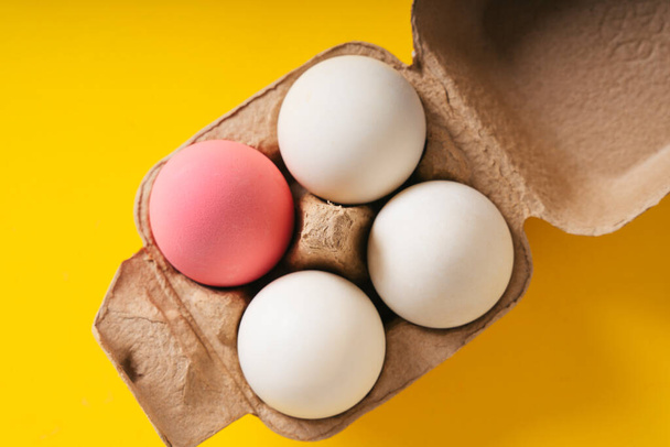 Draufsicht - Ein rosa Ei und drei weiße Eier in einer Schachtel vor gelbem Hintergrund. - Foto, Bild