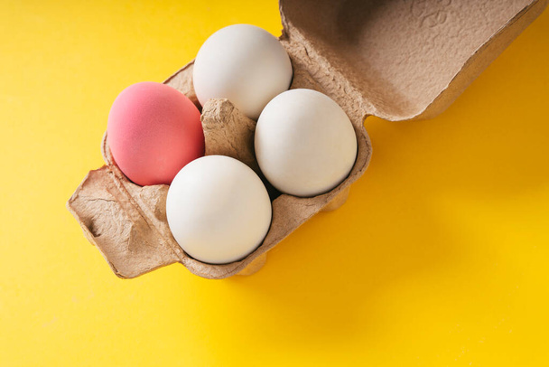 Vista superior: un huevo rosa y tres huevos blancos en una caja sobre fondo amarillo
. - Foto, Imagen