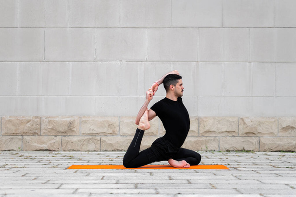 Hombre latino deportivo guapo haciendo ejercicio en una ciudad, haciendo yoga, entrenamiento de fitness o pilates, sentado en una pata King Pigeon Pose
. - Foto, Imagen