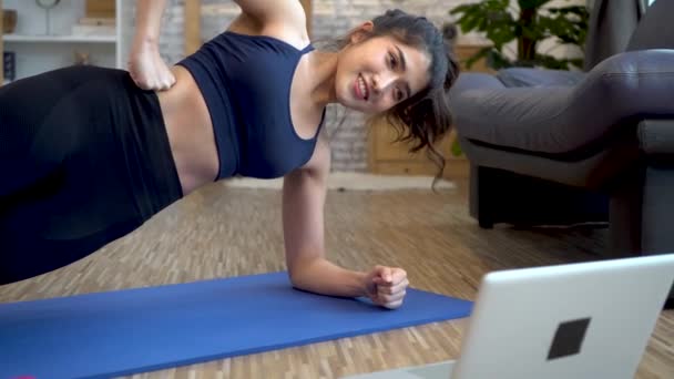 20s young Asian woman in sportswear doing stretching exercise while watching fitness training class on computer laptop online. Healthy girl exercising in living room with sofa couch in the background - 映像、動画