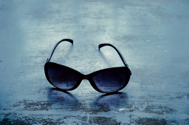 Blauwe moderne modieuze zonnebril op rustieke vloer. Mode achtergrond. Stillevens fotografie. Zonneschijn van achteren. Lensschaduw op de vloer. - Foto, afbeelding