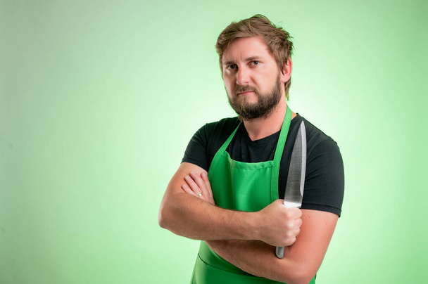 緑色のエプロンと黒のTシャツのヒーローを持つスーパーマーケットの従業員は、自信を持って緑の背景に隔離された彼の手にナイフを保持します - 写真・画像