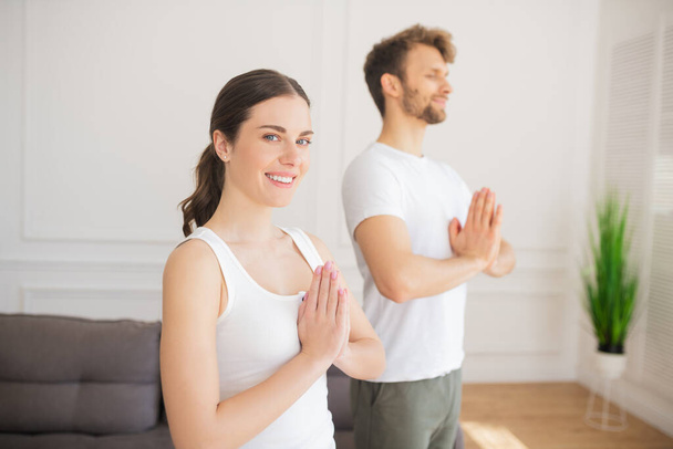 Young couple in white tshirts doing yoga at home putting their hands in namaste, woman smiling - Foto, afbeelding