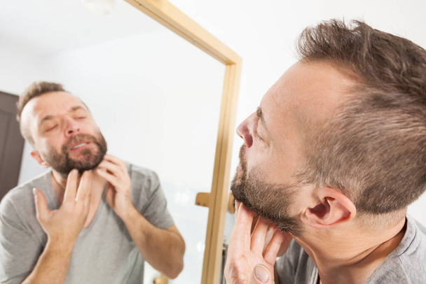 Mature adult man investigating his unshaved facial hair. Guy thinking about shaving his short beard - 写真・画像