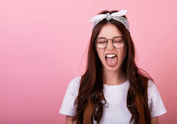 Dissatisfied young long-haired brunette woman hipster in a bandana, glasses, vest on a pink background. Lady emotionally grimaces and shows her tongue, not happy with the events. Copy space. - Foto, Bild