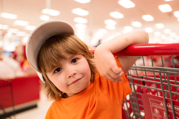 Little cute boy with shopping cart full of fresh organic vegetables and fruits standing in grocery department of food store or supermarket. - 写真・画像