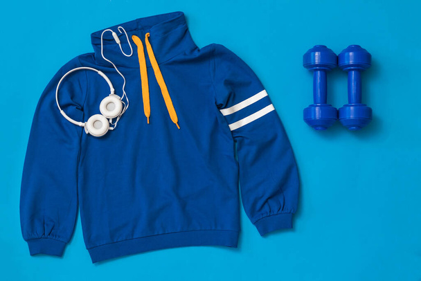 Пара синіх гантелів, навушників і спортивної куртки на синьому фоні
. - Фото, зображення