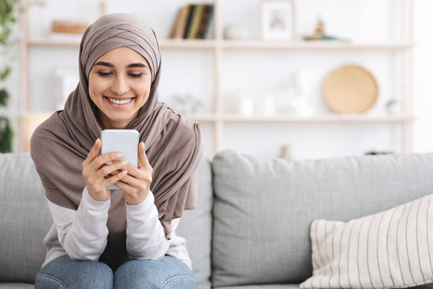 Χαρούμενη μουσουλμάνα κοπέλα με μαντίλα ξοδεύει χρόνο στο σπίτι με Smartphone - Φωτογραφία, εικόνα