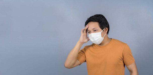Yüz maskesi takan Asyalı adam mavi arka planda baş ağrısı ve endişe, Coronavirüs salgını, covid19 salgını, sosyal uzaklık ve sorumluluk, sağlık ve virüs konseptine karşı koruma - Fotoğraf, Görsel