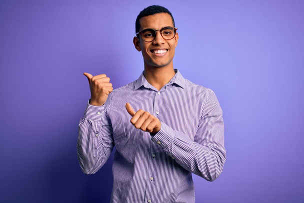 Handsome african american man wearing striped shirt and glasses over purple background Pointing to the back behind with hand and thumbs up, smiling confident - Foto, Imagen