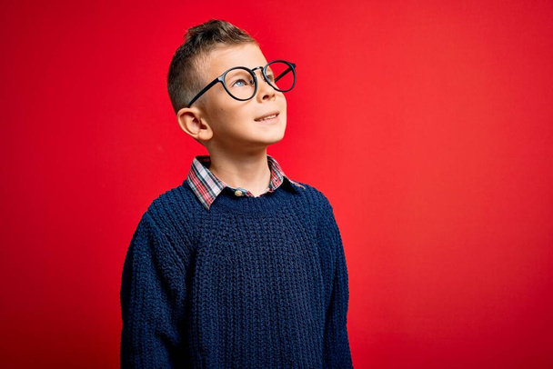 Junge kleine kaukasische Kinder mit blauen Augen stehen mit smarter Brille vor rotem Hintergrund und schauen mit einem Lächeln im Gesicht, natürlichem Gesichtsausdruck, zur Seite. Zuversichtliches Lachen. - Foto, Bild