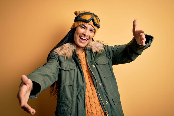 Mladá krásná hispánka v lyžařských brýlích a kabátě na zimní počasí, dívá se na kameru s otevřenou náručí a usmívá se. Veselý výraz objímající štěstí. - Fotografie, Obrázek