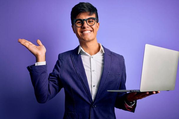Nuori komea liikemies silmälasien kanssa työskentelee kannettavan tietokoneen yli violetti tausta erittäin onnellinen ja innoissaan, voittaja ilme juhlii voittoa huutaen iso hymy ja nosti kädet - Valokuva, kuva