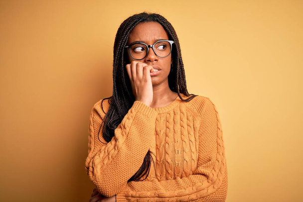 Молода афроамериканська розумна жінка в окулярах і повсякденному светрі на жовтому тлі виглядає напруженою і нервовою з руками на роті кусає цвяхи. Проблема тривоги
. - Фото, зображення