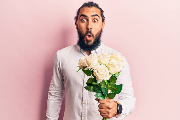 Jeune homme arabe tenant des fleurs effrayé et étonné avec la bouche ouverte pour la surprise, visage incrédule  - Photo, image
