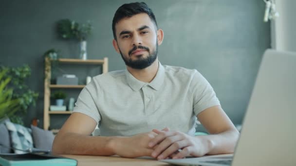 Portrait of attractive young Arab man looking at camera sitting at table with laptop at home - Filmagem, Vídeo