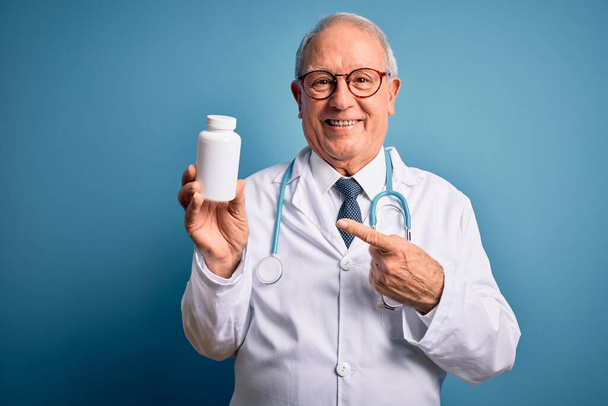 Senior gris doctor hombre sosteniendo píldoras farmacéuticas sobre fondo azul muy feliz señalando con la mano y el dedo
 - Foto, imagen