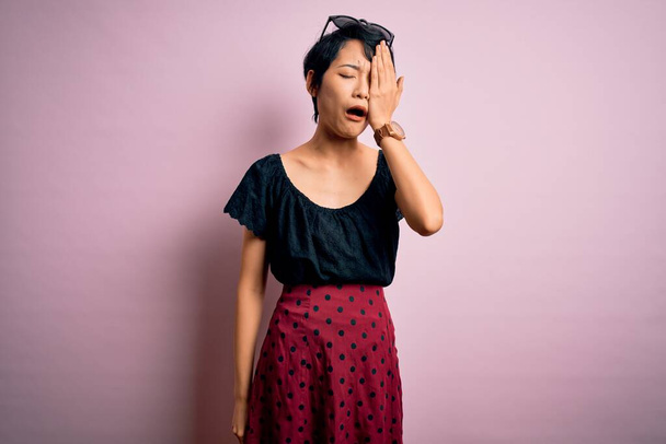 Junge schöne asiatische Mädchen in lässigem Kleid vor isoliertem rosa Hintergrund stehend Gähnend müde, das halbe Gesicht, Augen und Mund mit der Hand verdeckt. Gesicht schmerzt vor Schmerzen. - Foto, Bild