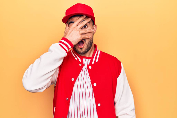 Handsome sporty man with beard wearing baseball jacket and cap over yellow background peeking in shock covering face and eyes with hand, looking through fingers afraid - 写真・画像