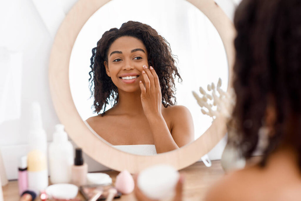 Pretty black woman looking in the mirror and applying facial moisturizer creme - Foto, Imagem