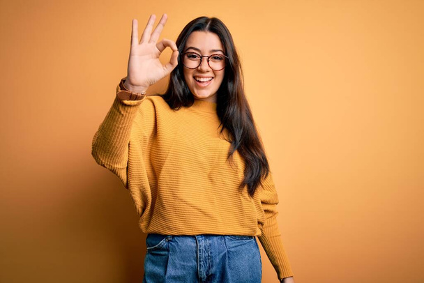 Young brunette woman wearing glasses and casual sweater over yellow isolated background smiling positive doing ok sign with hand and fingers. Successful expression. - Photo, Image