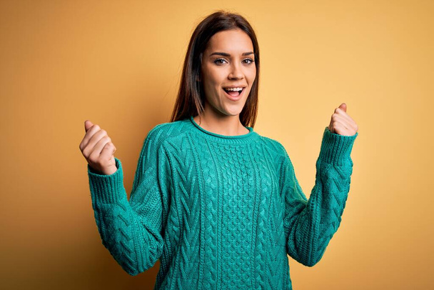 Junge schöne brünette Frau in grünem lässigen Pullover über gelbem Hintergrund feiert überrascht und erstaunt über den Erfolg mit erhobenen Armen und offenen Augen. Siegerkonzept. - Foto, Bild