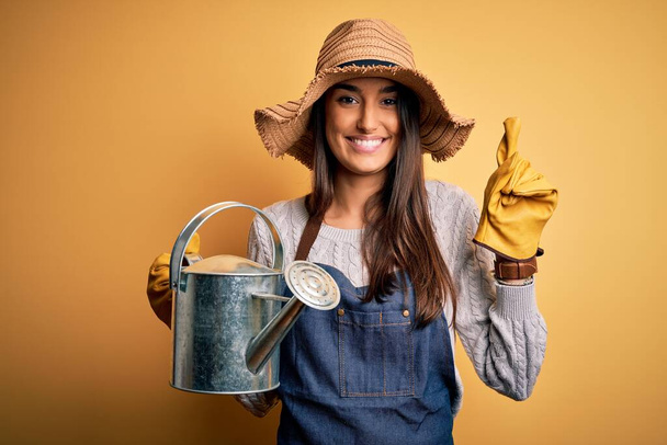 Νεαρή όμορφη μελαχρινή αγρότισσα γυναίκα φορώντας ποδιά και καπέλο κρατώντας πότισμα μπορεί να εκπλαγεί με μια ιδέα ή ερώτηση δείχνοντας δάχτυλο με χαρούμενο πρόσωπο, νούμερο ένα - Φωτογραφία, εικόνα