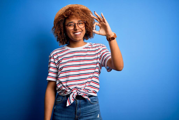 Afrikkalainen amerikkalainen nainen kihara tukka lomalla yllään kesähattu ja raidallinen t-paita hymyilevä positiivinen tekee ok merkki käsin ja sormin. Onnistunut ilmaisu. - Valokuva, kuva