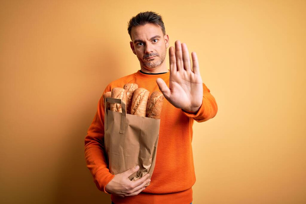 Молодий красивий чоловік тримає паперовий мішок з хлібом над ізольованим жовтим тлом з відкритою рукою робить знак зупинки з серйозним і впевненим виразом, жест захисту
 - Фото, зображення
